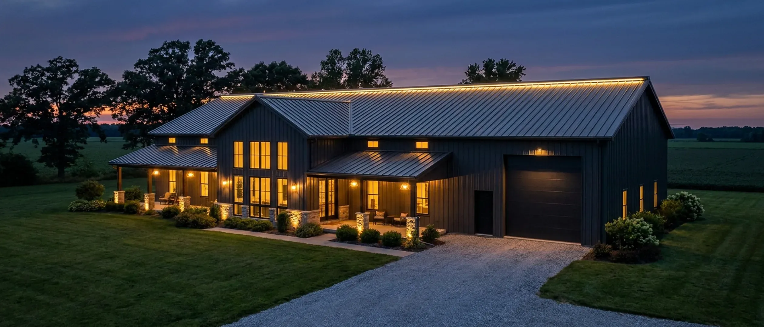 A barndominium at dusk with warm light glowing from every window and exterior lighting along the porch and roofline against a deep blue twilight sky.
