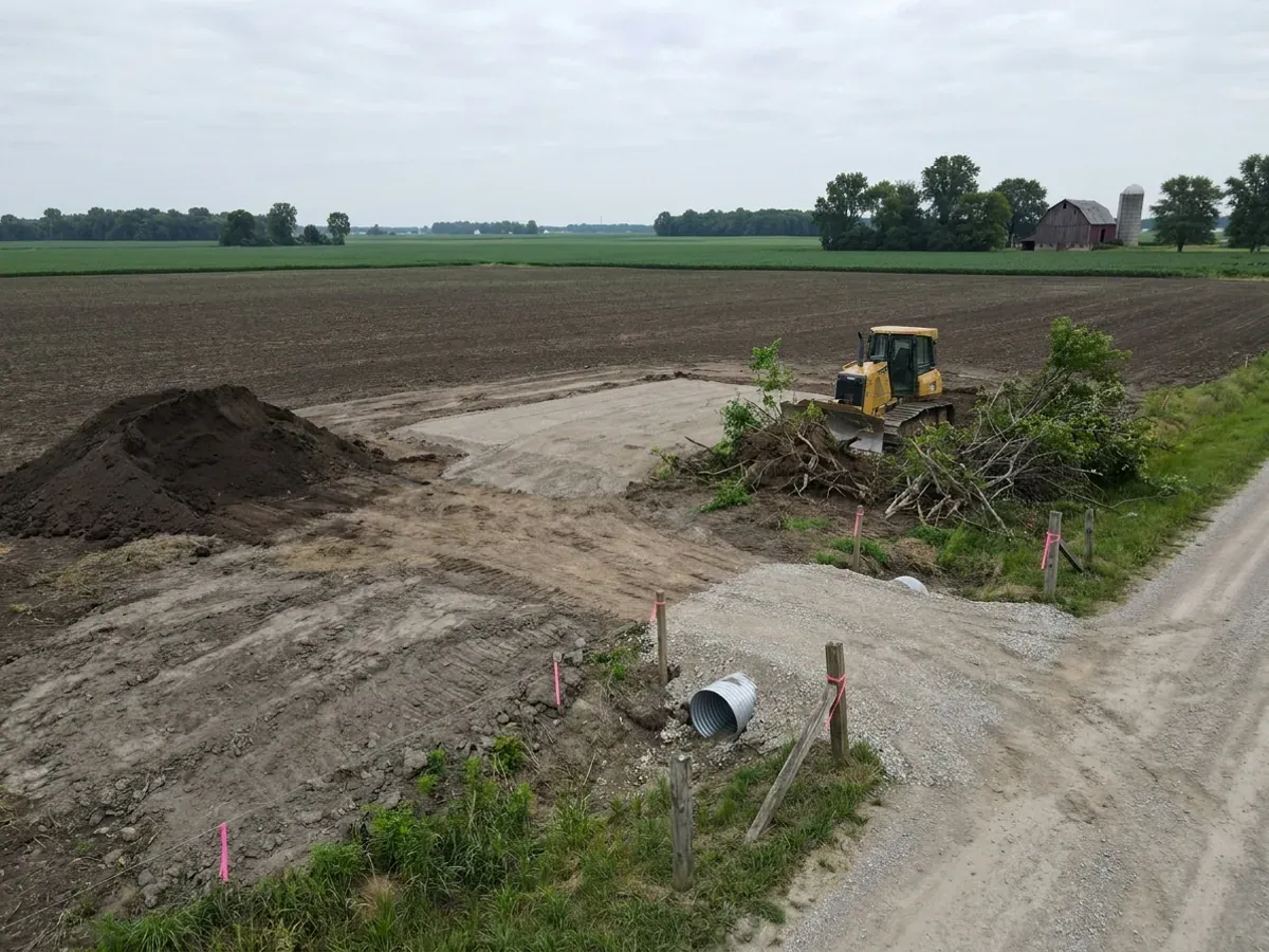 A dozer clearing brush on a level rural parcel beside a compacted crushed-stone building pad, with dark topsoil stripped back and a culvert set at the driveway entrance.