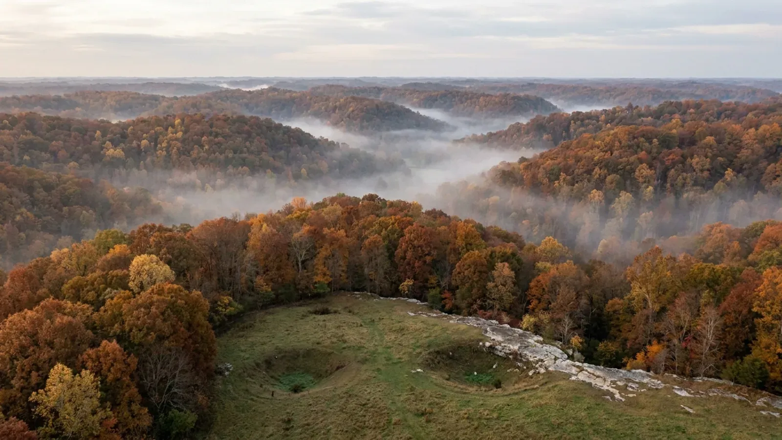 Steep forested hills in autumn colour with fog in the hollows, and a foreground pasture marked by grassy sinkholes and a pale limestone ledge.